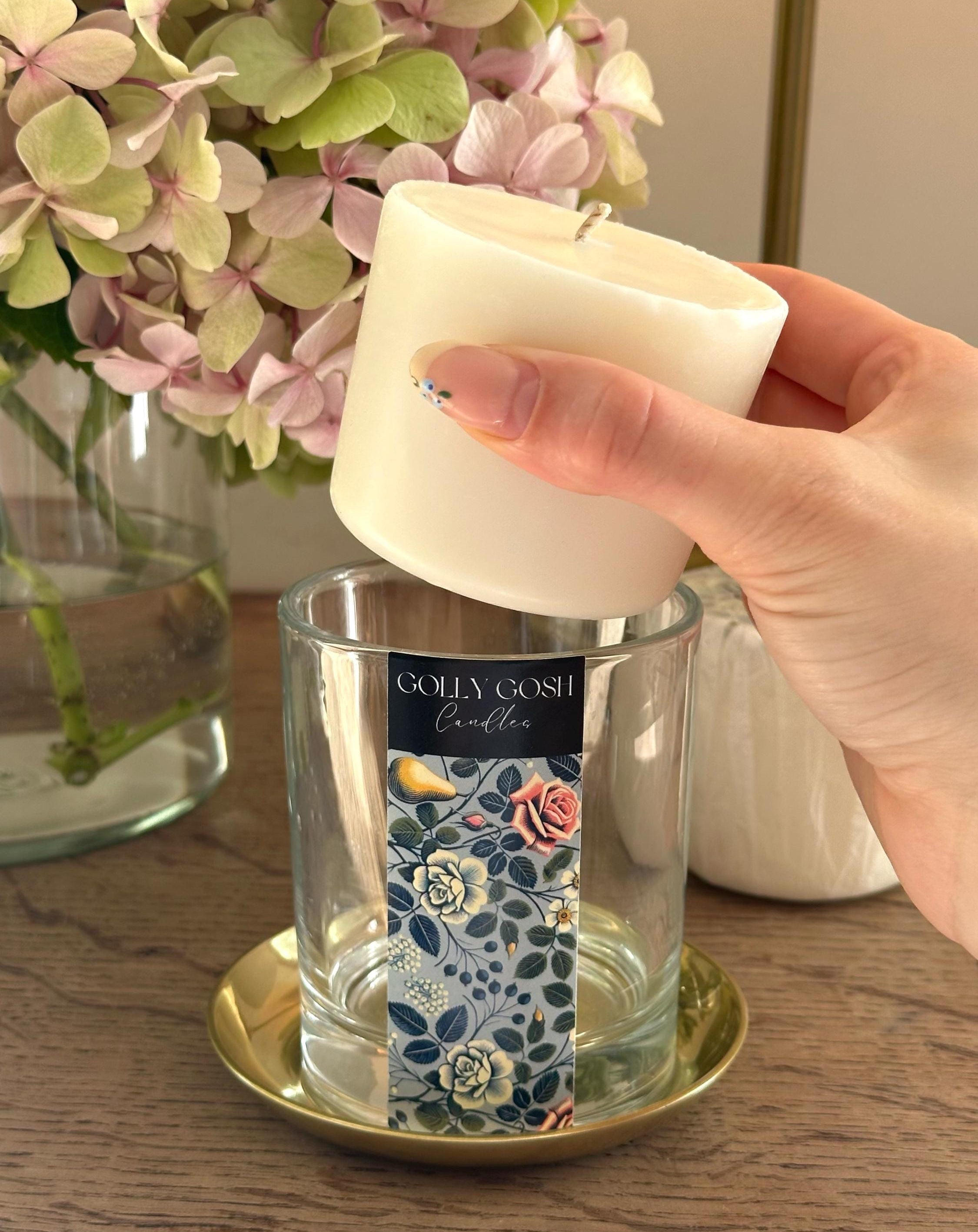 Hand placing a white candle into a glass holder with a floral label, surrounded by flowers and a vase.